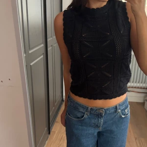 Svart stickad topp från Vero Moda - Svart stickad väst från Vero Moda med coolt virkat mönster och ribbade kanter. Modellen är ärmlös och croppad. Västen har en hög halsringning och ett unikt rutigt strukturmönster som ger en trendig vibe.