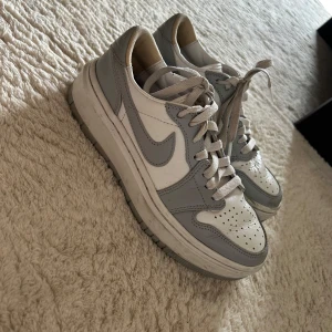 Nike Air Force 1 i vitt och grått - Snygga Nike Air Force 1 sneakers i vitt och ljusgrått skinn. Klassisk siluett med rund tå, perforerad tåbox och platt sula. Skorna har snörning och den ikoniska swoosh-loggan på sidorna. Perfekta för dig som gillar stilrena och tidlösa sneakers.