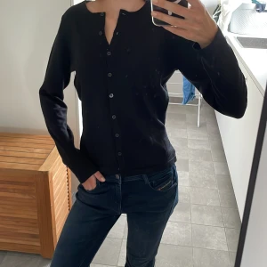 Svart kofta - En stilren svart kofta med långa ärmar och knappar hela vägen framtill. Koftan har en enkel, rak passform och är perfekt att slänga över en t-shirt eller linne. Materialet känns mjukt och lätt, vilket gör den skön att bära under kyligare dagar.