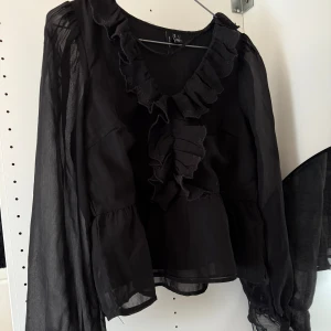 Svart blus med volangdetaljer - Säljer en svart blus med transparenta, långa ärmar och snygga volangdetaljer längs v-ringningen och framtill. Blusen har en luftig känsla och är perfekt för dig som gillar en romantisk och trendig stil. En liten fläck på framsidan men inget som märks💗💗