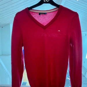 Röd v-ringad tröja J.Lindeberg  - Snygg röd v-ringad tröja från J.Lindeberg i storlek XS. Tröjan är stickad och har långa ärmar samt en liten broderad logga på bröstet. Perfekt för dig som gillar stilrena och klassiska plagg med en poppig färg.