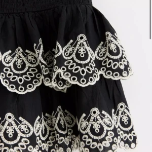 Svart volangkjol med broderi - Supersöt svart kortkjol med två volanger och vitt broderi längs kanterna. Kjolen har bred smock i midjan för en bekväm passform och ett unikt, detaljerat mönster som verkligen sticker ut. Perfekt för dig som vill ha något extra till outfiten. Knappt aldrig använd och ganska ny! 