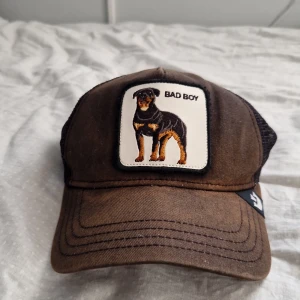Brun keps Goorin Bros Bad Boy - Snygg brun keps från Goorin Bros med mesh-bakstycke och justerbar snapback. Framtill finns ett broderat märke med en rottweiler och texten 'BAD BOY'. Perfekt accessoar för dig som vill sticka ut med en cool och unik look.