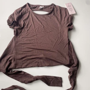 Brun omlottopp med knyt från Melly - Supersnygg brun omlottopp & kjol från Nelly med korta ärmar och långa knytband i midjan. Toppen har en rund halsringning och en öppen detalj i ryggen. 