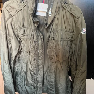Olivgrön vindjacka från Moncler unisex - Snygg olivgrön vindjacka från Moncler med klassisk logga på ärmen. Jackan har flera fickor med lock, axelklaffar och både dragkedja och knappar framtill. Tillverkad i lätt polyester och har lång ärm med knappdetalj vid mudden. Passar både kvinna och man då både jag och min bror haft den. Storlek 2 motsvarande M. Gott skick.