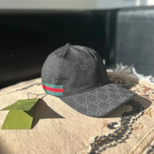Svart Gucci keps med GG-mönster - Snygg svart keps från Gucci med klassiskt GG-monogram över hela kepsen. På sidan finns en ikonisk grön och röd rand. Kepsen har böjd skärm och är tillverkad i ett slitstarkt tygmaterial. Perfekt för dig som vill ha en stilren och lyxig accessoar.