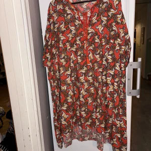 Röd mönstrad kortklänning med snörning - Säljer en röd kortklänning med färgstarkt paisleymönster i vitt, gult och svart. Klänningen har korta ärmar, v-ringning med snörningsdetalj och ett luftigt, flowy tyg som känns perfekt för varma dagar. Superfin till sneakers eller sandaler!