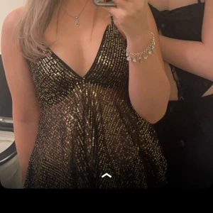 Glittrig topp - Säljer en glittrig svart klänning med guldiga paljetter och v-ringning. Klänningen har tunna axelband och ett flowy fall, perfekt för att sticka ut. Materialet känns lätt och skimrande, vilket ger en festlig look.
