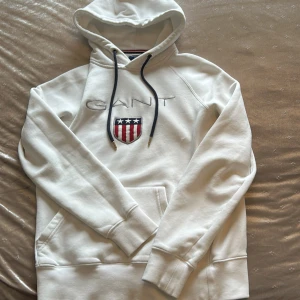 Vit hoodie från GANT med broderad logo - Vit hoodie från GANT med broderad logo och sköld på bröstet. Tröjan har huva med mörkblå snören, känguruficka och långa ärmar. Klassisk och stilren design, perfekt för dig som gillar preppy vibes.