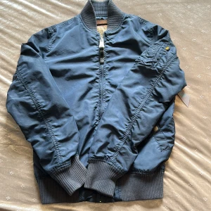 Blå bomberjacka Alpha Industries S - Säljer en klassisk blå bomberjacka från Alpha Industries med orange foder. Jackan har ribbade muddar, dragkedja framtill och en ikonisk ficka med dragkedja på ärmen. Tillverkad i slitstark polyester och har en snygg, tidlös look.