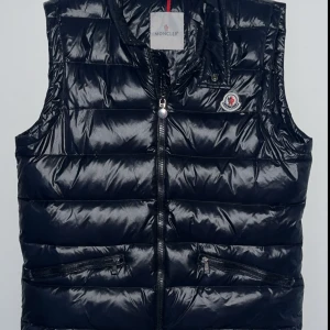Svart dunväst från Moncler - Snygg svart dunväst märke Moncler med glansig finish och klassisk logga på bröstet. Västen har två dragkedjefickor framtill och stängs med dragkedja. Perfekt att slänga över en hoodie för en cool look. Använd 2 gånger.