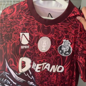 FC Porto röd matchtröja med drakmönster - Snygg FC Porto fotbollströja från New Balance i rött med mörkröda och svarta drakmönster. Kortärmad modell med klubbmärke, sponsorloggor och unikt tryck över hela tröjan. Perfekt för dig som vill sticka ut på läktaren eller planen.