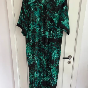 Klänning / Kaftan  - Säljer en långklänning från Lindex med grönt och svart bladmönster. Klänningen har 3/4 ärmar, knytning vid halsen och en loose passform. Perfekt för dig som gillar statement-plagg med coolt mönster och avslappnad stil.