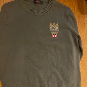Olivgrön stickad tröja från Morris - Snygg olivgrön stickad tröja från Morris med broderad logga och brittisk flagga på bröstet. Storlek S. Kan gå ner i pris för smidig affär.