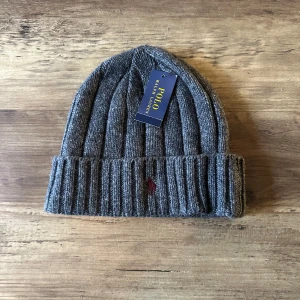 Grå ribbad mössa från Polo Ralph Lauren OANVÄND - Snygg grå ribbstickad mössa från Polo Ralph Lauren med uppvikt kant och diskret broderad logga framtill. Perfekt accessoar för kyliga dagar och enkel att matcha med olika outfits.