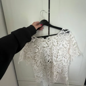Vit blommig spetsblus - Superfin vit blus i spets med stora blommiga mönster och korta ärmar. Luftig och genombruten design som ger en romantisk vibe. Perfekt att styla över ett linne eller bikini för en cool look. Känns som en S men står L i lappen 