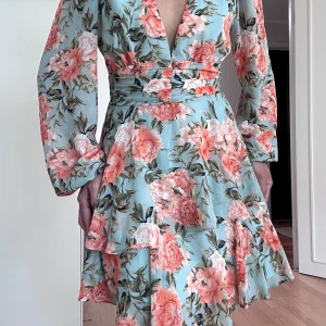 Brand new summer dress från Hello Molly - Supersöt blommig kortklänning från Hello Molly i ljusblått med stora rosa och korallfärgade blommor. Klänningen har v-ringning, långa ärmar och volangdetaljer i kjolen. Perfekt för sommarens alla tillfällen!