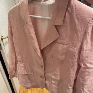 Ljusrosa kavaj i linne - Snygg ljusrosa kavaj med klassisk krage och tre fickor framtill. Kavajen har två knappar och är tillverkad i ett luftigt linnematerial, perfekt för vår och sommar. Passformen är normal och ärmarna är långa. 