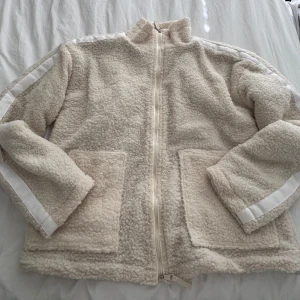 Beige teddyjacka med dragkedja - Mysig beige teddyjacka med hel dragkedja framtill och två stora fickor. Jackan har vita ränder längs ärmarna och en hög krage som ger extra stilpoäng. Perfekt för kyliga dagar när du vill vara både varm och snygg.