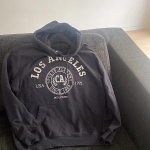 Svart hoodie Los Angeles tryck - Svart hoodie med stor vit Los Angeles-print framtill och sportig collegekänsla. Klassisk känguruficka, snörning i huvan och mjuk insida. Perfekt för chill dagar eller när du vill ha en avslappnad streetstyle.