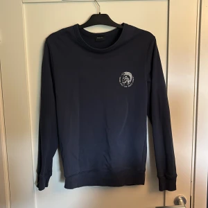 Mörkblå sweatshirt från Diesel - Snygg mörkblå sweatshirt från Diesel med klassiskt tryck på bröstet. Tröjan har rund hals och långa ärmar. Perfekt för en chill och stilren look. Passar dig som gillar streetwear och vill ha något bekvämt men ändå trendigt.