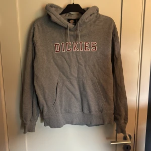 Grå hoodie från Dickies - Säljer en grå hoodie från Dickies i storlek medium. Tröjan har huva med snörning, stor magficka och Dickies-logga i rött och vitt på bröstet. Lite uttvättad men annars i fint skick. Perfekt för chill dagar.