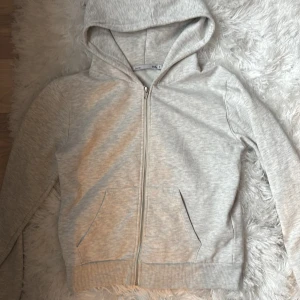 Ljusgrå hoodie med dragkedja - En stilren ljusgrå hoodie med dragkedja och huva. Tröjan har två fickor framtill och ribbade muddar vid ärmslut och nederkant. Perfekt för en avslappnad look och enkel att matcha med jeans eller mjukisbyxor.