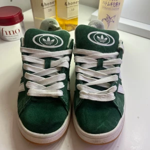 Adidas Campus gröna  - Gröna Adidas Campus sneakers i mocka med vita detaljer och klassiska tre ränder på sidan. Skorna har vit sula, vita skosnören och låg profil. Perfekta för dig som gillar retrostil och streetwear. Köpt för 1000 och är nytvättade. Man får med helt oanvända gröna snören också. 