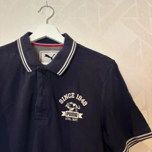 Mörkblå pikétröja från Puma - Snygg mörkblå pikétröja från Puma med vit broderad logga och text 'Since 1948' på bröstet. Storlek S