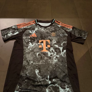 FC Bayern München Adidas fotbollströja - Säljer en FC Bayern München fotbollströja från Adidas med svart och grått marmorerat mönster, orange detaljer och loggor. Tröjan har korta ärmar, klassiska Adidas-ränder på axlarna och är tillverkad i lätt funktionsmaterial (Aeroready).