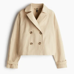 Beige trenchcoat med knappar - Stilren och kort trenchcoat i beige med dubbelknäppning framtill och klassisk krage. Jackan har långa ärmar med snygga detaljer vid ärmsluten och är tillverkad i ett mjukt bomullsmaterial. Perfekt för dig som gillar en clean och tidlös look. Slutsåld i alla storlekar❤️