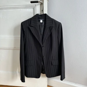 Pinstripe kavaj - Snygg svart kavaj med tunna vita ränder. Klassisk modell med två knappar framtill, breda slag och fickor med lock. Från tidigt 2000-tal men i fint skick! 