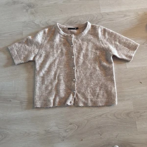 Beige kofta med pärlknappar från 157 - Superfin beige kofta från 157 i stickad kvalitet. Kortärmad modell med rund hals och snygga pärlknappar framtill. Perfekt att slänga över en topp för en chill och stilren look. Ribbstickade kanter ger extra detalj.