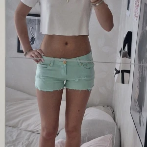 Lågmidjade färgglada shorts - Snygga och trendiga mintgröna jeansshorts från Madonna med slitna detaljer och råa kanter. Klassisk femficksmodell med gylf och knapp, samt bälteshällor. Perfekta för varma dagar när du vill sticka ut lite extra.
