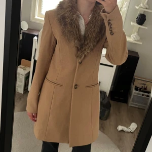 Beige kappa med fuskpäls-krage - Snygg beige kappa med markerad midja och stor krage i fuskpäls. Kappan har knappar vid ärmslut och en enkel knäppning framtill. Perfekt för dig som vill ha en klassisk men trendig look under höst och vinter. Passar mig som brukar ha xs/s