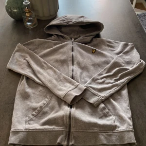 Lyle and scott hoodie - Snygg grå hoodie från Lyle & Scott med dragkedja och huva. Klassisk gul logga på bröstet och två fickor framtill. Perfekt för chill dagar eller när du vill ha en enkel och stilren look.
