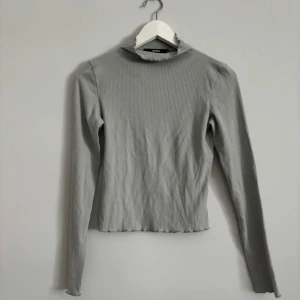Ljusgrå ribbad polotröja Bik Bok - Supersnygg ljusgrå ribbad polotröja från Bik Bok i storlek S. Tröjan har lång ärm, lätt croppad passform och en liten volangkant vid halsen. Perfekt att styla med jeans eller kjol för en clean look.