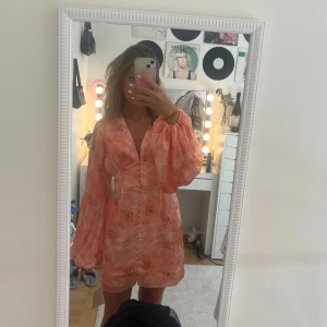Peachfärgad blommig kortklänning från Adoore - Superfin kort klänning från Adoore i peach och rosa toner med blommigt mönster. Djup v-ringning, långa puffärmar och klädda knappar framtill. Sitter snyggt i midjan och har ett lätt glansigt material. Helt ny med prislapp kvar!