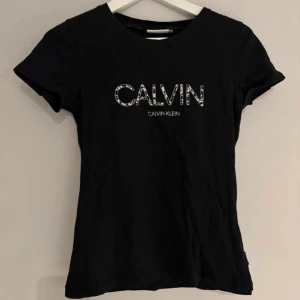 Svart Calvin Klein t-shirt med logga - Svart t-shirt från Calvin Klein med korta ärmar och rund hals. Framsidan har en stor CALVIN-logga i vitt med mönstrad kant och mindre Calvin Klein-tryck under. Skön passform i mjuk bomull, perfekt till jeans eller shorts.