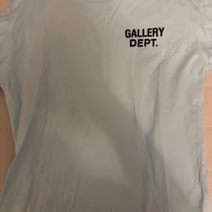 Ljusblå t-shirt från Gallery Dept. - Snygg ljusblå t-shirt från Gallery Dept. med svart logga tryckt på bröstet. Klassisk rund hals och korta ärmar, perfekt för en chill och stilren look. Tillverkad i mjuk bomull som känns skön mot huden.