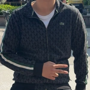 Svart mönstrad vindjacka från Lacoste och byxor  - Säljer en svart vindjacka från Lacoste med diskret geometriskt mönster och gröna samt vita ränder längs ärmarna. Jackan har dragkedja framtill, ståkrage och den klassiska Lacoste-loggan på bröstet. Perfekt för dig som gillar sportig och clean stil.