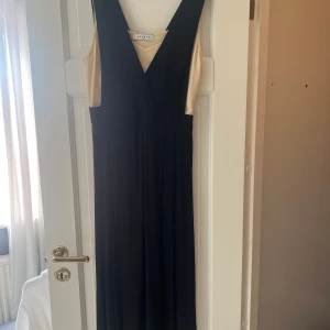 Svart mellanlång klänning från Sandro Paris - Elegant svart långklänning från Sandro Paris med djup v-ringning och ärmlös design. Klänningen har en snygg kontrast med ljus, champagnefärgad insida och ett mjukt, flytande tyg som ger en stilren siluett. Perfekt för dig som vill sticka ut med klassisk stil.