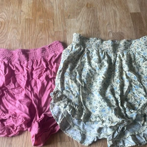 2-pack shorts, blommiga & rosa, CUBUS - Två par shorts från Cubus. Ena paret är ljusbeige med blått blommönster, volangdetaljer och smock i midjan. Det andra paret är helrosa med smockad midja och lös passform. Perfekta för sommaren och riktigt sköna att ha på sig.45:-st