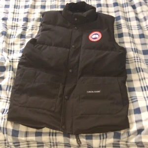 Svart dunväst från Canada Goose . KAN SÄNKA VID SNABB AFFÄR - Svart dunväst från Canada Goose med klassisk logga på bröstet och stora fickor framtill. Västen har hög krage, tryckknappar och dragkedja. Perfekt för lager-på-lager och håller dig varm under kyliga dagar. Passar folk som är 160-170 cm. Dragkejde lappen är lite trasig men inget som syns och den funkar bra