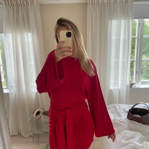 Röd kortklänning med knyt i midjan - Snygg röd kortklänning från Gina Tricot med vida långa ärmar och knytband i midjan. Klänningen har en enkel, stilren design och är gjord i ett mjukt, lätt material som känns skönt mot huden. Perfekt för dig som vill sticka ut med färg och cool passform.