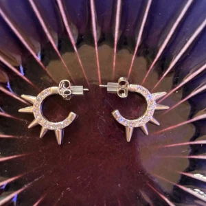 Silvriga örhängen med spikes och stenar - Snygga Coola Stockholms stills örhängen slö e silvriga  i halvcirkel med gnistrande stenar och vassa spikes runt om. Perfekta för dig som vill sticka ut och addera lite edge till din stil. Små men iögonfallande, passar både till vardagslooken och när du vill glänsa extra. Helt nya aldrig andvända 