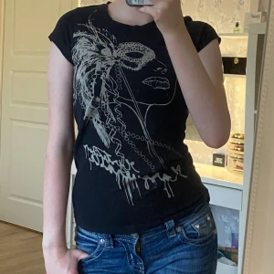 Svart t-shirt med maskmotiv Gina Tricot - Svart t-shirt från Gina Tricot med coolt tryck av ett ansikte med mask och fjädrar i silver. Modellen är figurnära med korta ärmar och rund hals. Perfekt för dig som gillar unika prints och vill sticka ut lite extra.