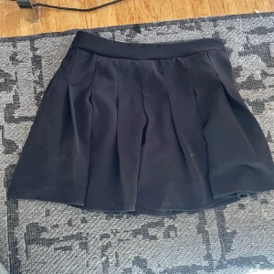 Svart plisserad kjol från SHEIN - Svart kortkjol från SHEIN i storlek M med snygga plisserade veck och bred linning. Kjolen har dragkedja i sidan för enkel på- och avtagning. Perfekt för dig som gillar en klassisk och stilren look.