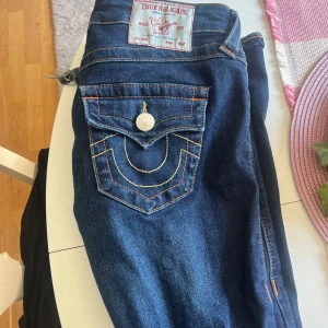 True Religion Joey Low Rise Flare Jeans - Mörkblå jeans från True Religion, modell Joey Low Rise Flare. Snygga kontrastsömmar, ikonisk bakficka med lock och knapp samt klassisk True Religion-lapp bak. Jeansen har låg midja och är flare. Lite slitet som man ser på bild 3 men annars bra skick!!