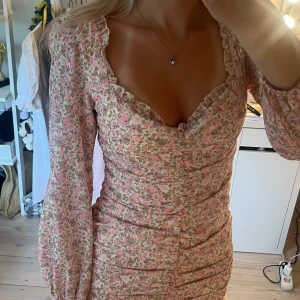  kortklänning från Nelly  - Supersöt kortklänning med rosa blommönster, volangkant nertill och vid urringning. Långa puffärmar med resår vid handlederna och smickrande rynkdetaljer längs hela klänningen. Perfekt för sommaren 